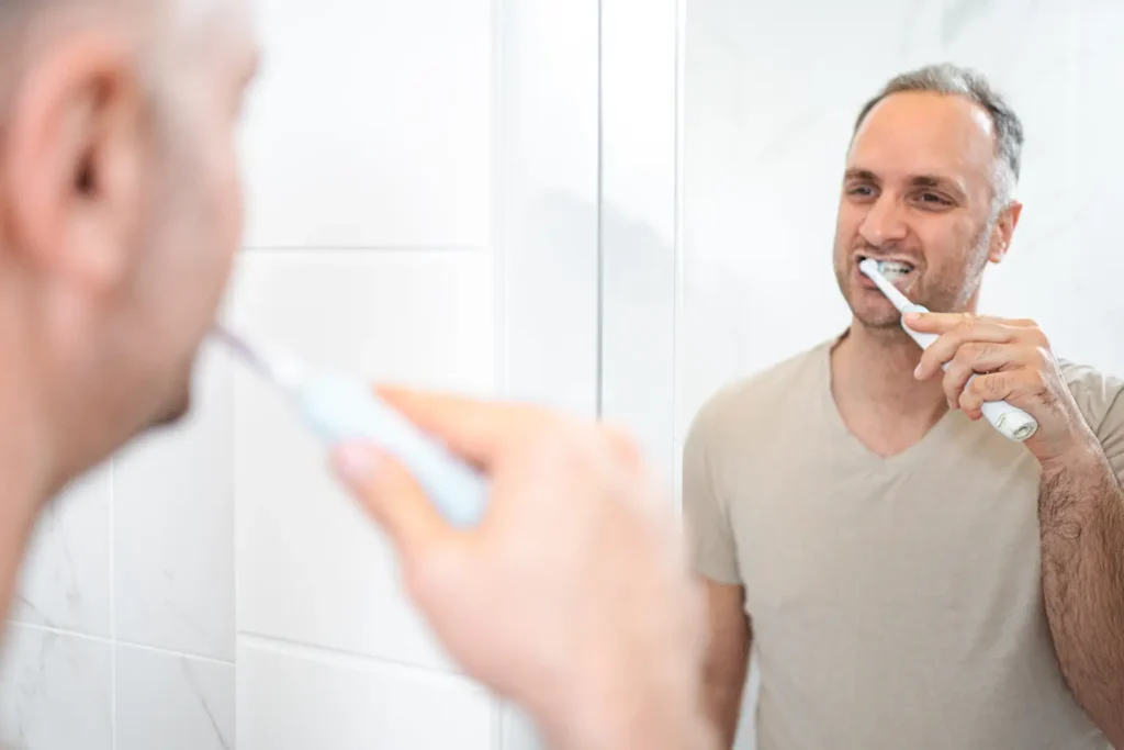 A man is brushing his teeth with an electric toothbrush in a bright, white-tiled bathroom. The image promotes the message "transform your smile" with the help of Big Sky Family Dental. The focus is on maintaining a healthy oral hygiene routine for a radiant and confident smile.