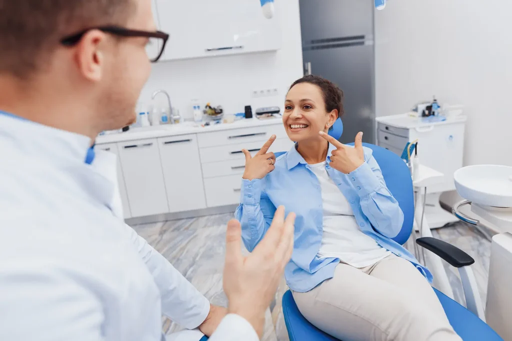 Cheerful woman pointing at her healthy teeth while discussing with a male dentist at Big Sky Family Dental. Learn how to maintain oral health despite the challenges of allergies.