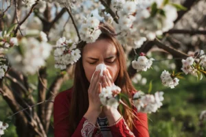 Woman experiencing allergies, blowing her nose surrounded by blooming flowers. Understanding the impact of allergies on oral health. Big Sky Family Dental provides insights on maintaining dental health during allergy season