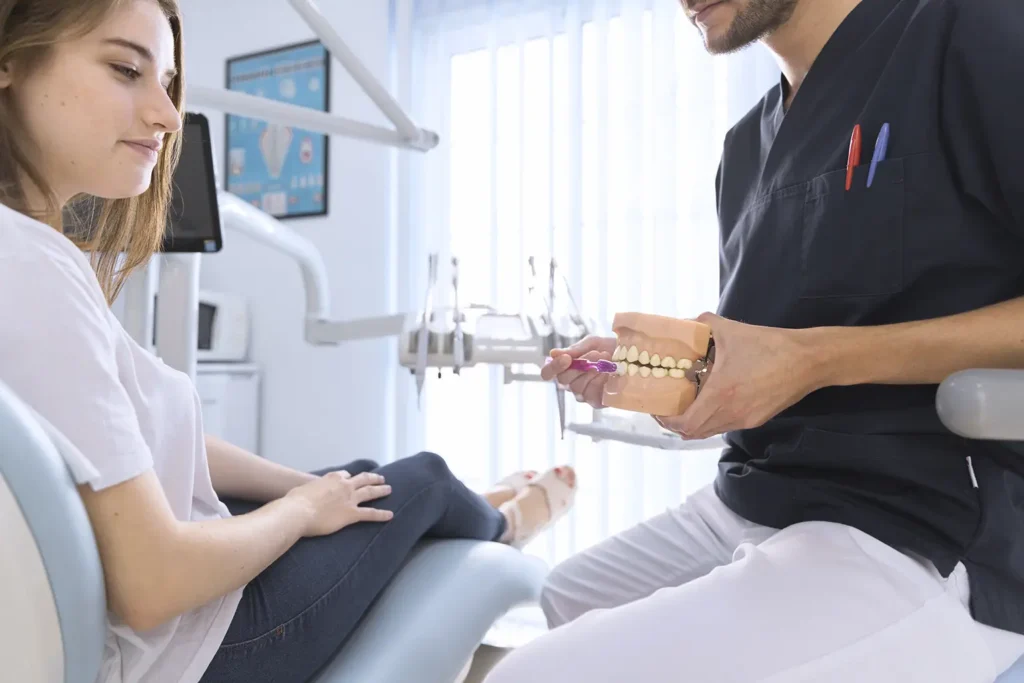 A dentist at Big Sky Family Dental in Billings, MT demonstrates proper brushing techniques to a patient using a teeth model. The patient appears to be attentive, possibly addressing her concern of "my teeth hurt." This instructional moment aims to improve dental hygiene and alleviate tooth pain.