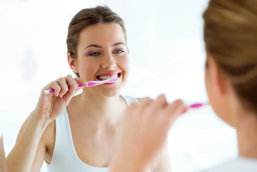 A young woman is brushing her teeth in front of a bathroom mirror, smiling as she practices good oral hygiene. This image highlights the importance of addressing concerns such as "my teeth hurt" and "teeth sensitivity." For personalized dental care, Big Sky Family Dental in Billings, MT offers expert solutions to maintain a healthy smile.